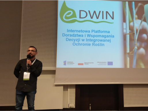 Maciej Zacharczuk stoi i wygłasza wykład. W tle telebim, na którym widać logo projektu eDWIN.